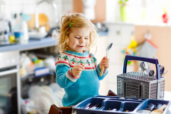 Jolie petite fille tout-petit aidant dans la cuisine avec lave-vaisselle. Joyeux enfant blond sain triage couteaux, fourchettes, cuillères, couverts. Bébé s'amuser à aider les travaux ménagers mère et père. — Photo