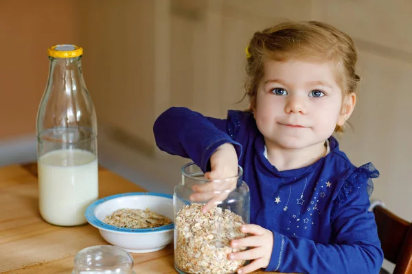Schattig peutermeisje dat gezonde havermout eet met melk als ontbijt. Schattig gelukkig baby kind in kleurrijke kleren zitten in de keuken en plezier hebben met het bereiden van haver, granen. Binnenshuis thuis — Stockfoto