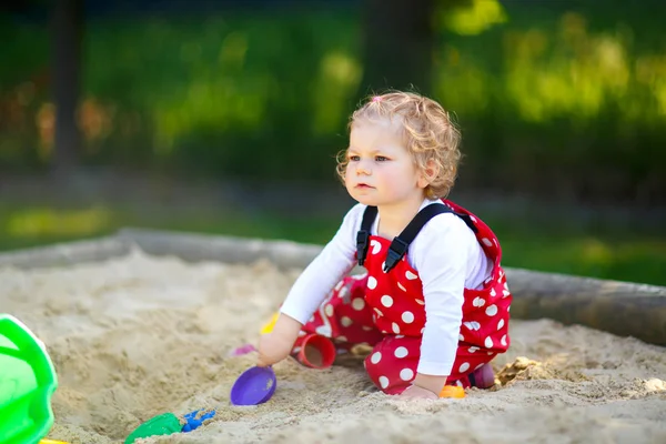 Söt liten flicka leker i sand på utomhus lekplats. Vacker baby i röda tuggummibyxor som har kul på solig varm sommardag. Barn med färgglada sandleksaker. Friska aktiva barn utomhus spelar spel — Stockfoto