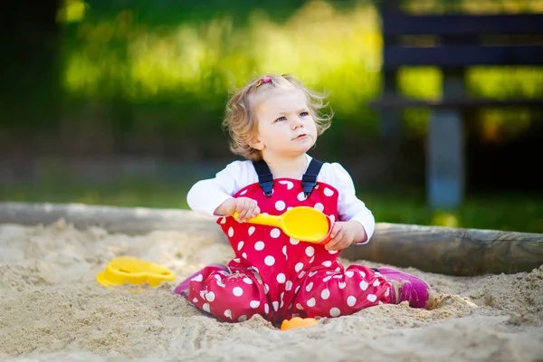 Schattig peuter meisje spelen in zand op buiten speeltuin. Mooie baby in rode gom broek die plezier heeft op zonnige warme zomerdag. Kind met kleurrijk zandspeelgoed. Gezonde actieve baby buiten speelt spelletjes — Stockfoto