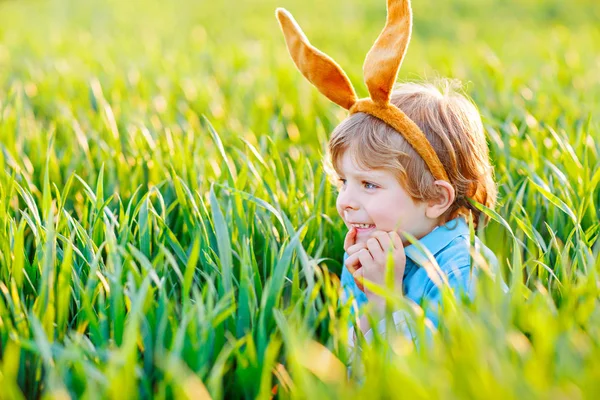 Ragazzino carino con orecchie da coniglio che si diverte con le tradizionali uova di Pasqua a caccia nelle calde giornate di sole, all'aperto. Festeggiamo la festa di Pasqua. Trovare bambino, uova colorate in erba verde — Foto Stock