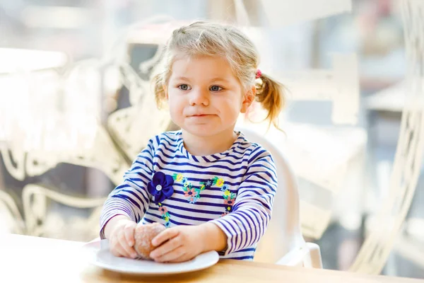 Linda niñita adorable sentada en el restaurante de la cafetería interior. Feliz niño sano comiendo pan o bollo dulce en un día soleado . —  Fotos de Stock