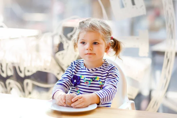 Linda niñita adorable sentada en el restaurante de la cafetería interior. Feliz niño sano comiendo pan o bollo dulce en un día soleado . —  Fotos de Stock