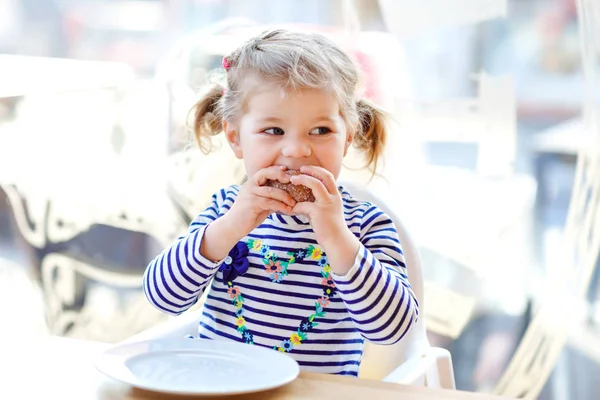 Cute little adorable toddler girl sitting in indoor cafe restaurant. Happy healthy baby child eating bread or sweet scone on sunny day. — Stock Photo, Image