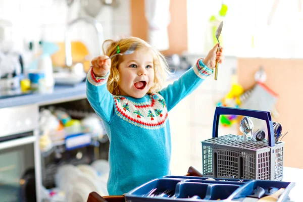 Aranyos kislány segít a konyhában mosogatógéppel. Boldog egészséges szőke gyerek válogató kések, villák, kanalakat, evőeszközök. Baby szórakozás segítésével házimunka anya és apa. — Stock Fotó