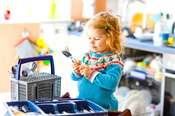 Küçük tatlı kız mutfakta bulaşık makinesiyle yardım ediyor. Bıçakları, çatalları, kaşıkları, çatalları ayıran mutlu sarışın çocuk. Bebek anne ve babaya yardım ederken eğleniyor.. — Stok fotoğraf