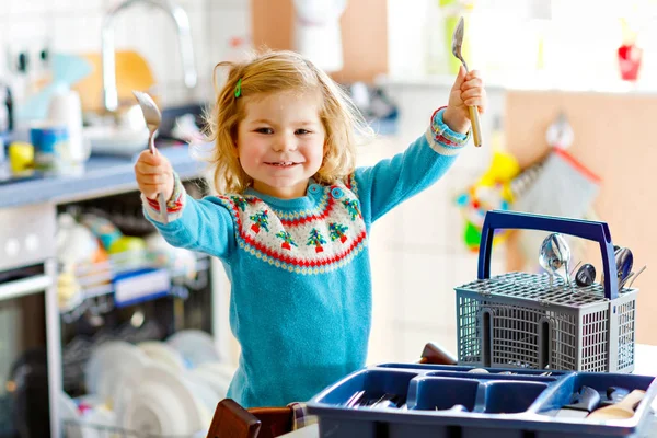 Linda niña pequeña ayudando en la cocina con lavavajillas lavadora. Feliz niño rubio sano clasificando cuchillos, tenedores, cucharas, cubiertos. Bebé divertirse con ayudar a las tareas domésticas madre y padre. —  Fotos de Stock