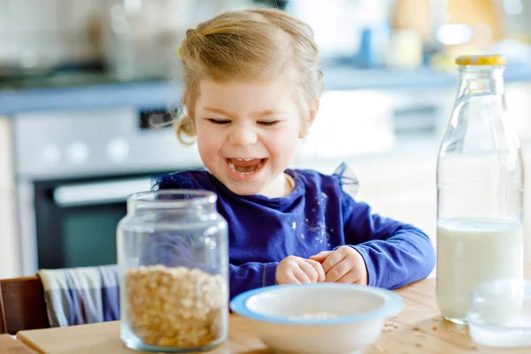 Entzückendes Kleinkind, das gesunde Haferflocken mit Milch zum Frühstück isst. Nettes glückliches Baby in bunten Kleidern sitzt in der Küche und hat Spaß mit der Zubereitung von Hafer, Getreide. Zuhause drinnen — Stockfoto
