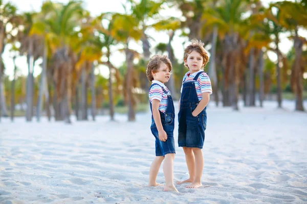 Due bambini piccoli ragazzi che si divertono sulla spiaggia tropicale, felici migliori amici che giocano, concetto di amicizia. Fratelli fratelli, gemelli in famiglia guardano con palme alberi su sfondo. Vacanze in famiglia. — Foto Stock