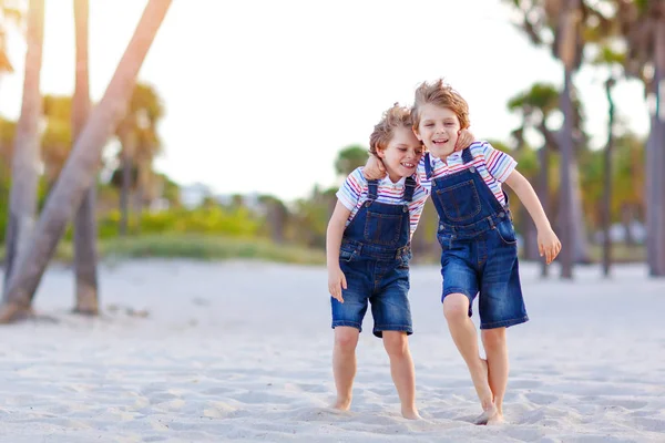 Two little kids boys having fun on tropical beach, happy best friends playing, friendship concept. Siblings brothers, twins fighting, running and jumping in family look with palms trees on background. — Stock Photo, Image