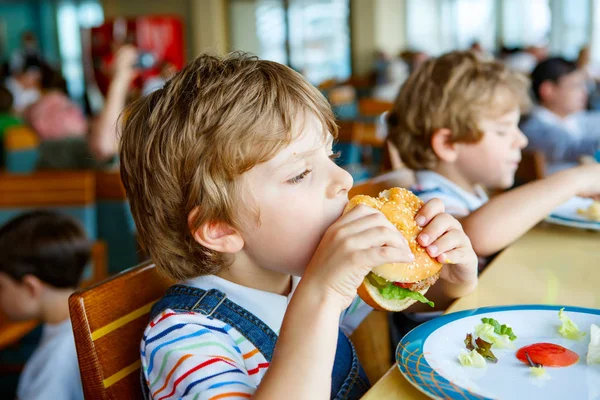 Милий здоровий дошкільний хлопчик їсть гамбургер, сидячи в шкільній їдальні — стокове фото