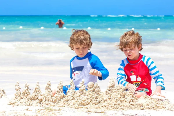 Dos niños divirtiéndose con la construcción de un castillo de arena en la playa tropical en la isla. Niños sanos jugando juntos en sus vacaciones. Gemelos, hermanos felices riendo y sonriendo — Foto de Stock
