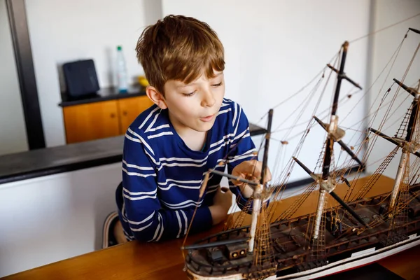 Kleiner blonder Schuljunge, der drinnen mit einem Segelschiff-Modell spielt. aufgeregtes Kind mit Jacht, das nach der Schule zu Hause Spaß hat. Glücklicher Junge baut und bastelt Spielzeug — Stockfoto