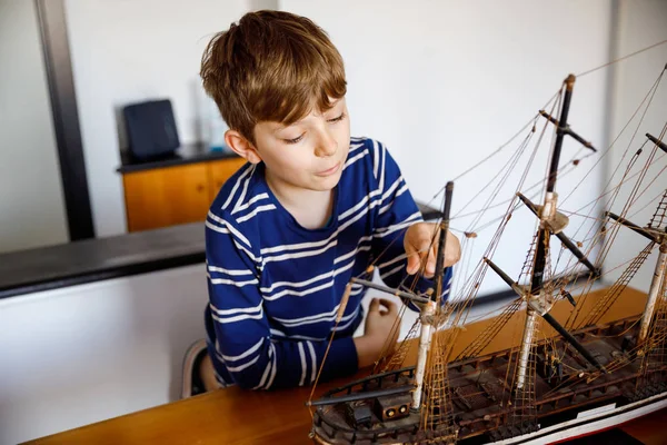 Lilla blonda skolan unge pojke leker med segelfartyg modell inomhus. Upphetsad barn med Yacht ha roligt efter skolan hemma. Happy Boy Building och skapa leksaker — Stockfoto