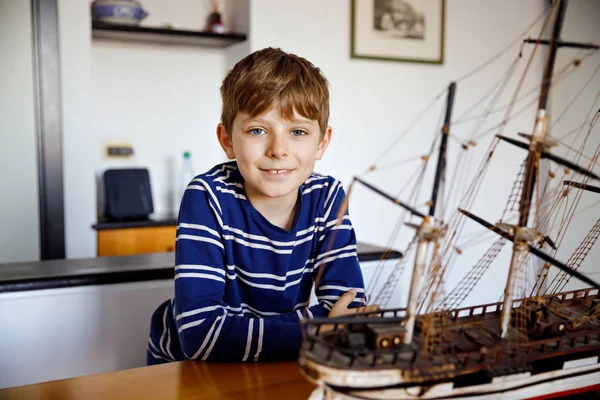 Petit écolier blond jouant avec le modèle de voilier à l'intérieur. Enfant excité avec yacht s'amuser après l'école à la maison. Joyeux garçon construire et créer des jouets — Photo