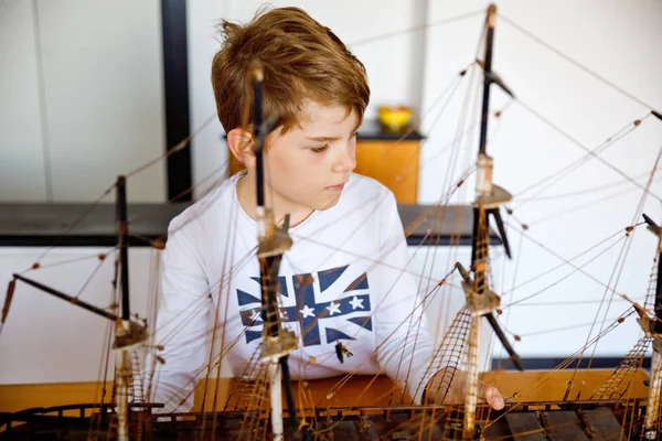 Kleiner blonder Schuljunge, der drinnen mit einem Segelschiff-Modell spielt. aufgeregtes Kind mit Jacht, das nach der Schule zu Hause Spaß hat. Glücklicher Junge baut und bastelt Spielzeug — Stockfoto