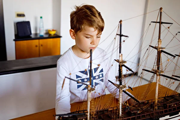 Pequeño niño de escuela rubia jugando con el modelo de barco de vela en el interior. Niño emocionado con yate divirtiéndose después de la escuela en casa. Feliz niño construyendo y creando juguetes — Foto de Stock