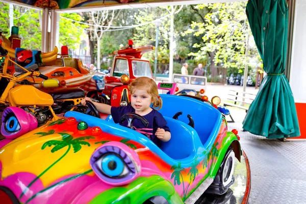 Adorable little toddler girl riding on funny car on roundabout carousel in amusement park. Happy healthy baby child having fun outdoors on sunny day. Family weekend or vacations — Stock Photo, Image