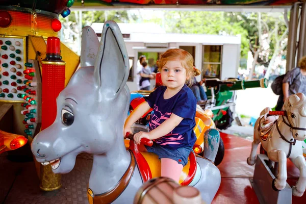 Adorable niña pequeña montado en animal en carrusel rotonda en el parque de atracciones. Feliz bebé sano que se divierte al aire libre en el día soleado. Fin de semana familiar o vacaciones — Foto de Stock