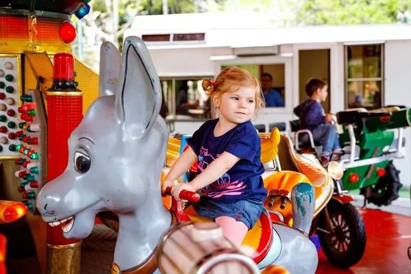 Adorable little toddler girl riding on animal on roundabout carousel in amusement park. Happy healthy baby child having fun outdoors on sunny day. Family weekend or vacations — Stock Photo, Image