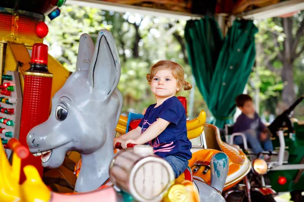 Adorable little toddler girl riding on animal on roundabout carousel in amusement park. Happy healthy baby child having fun outdoors on sunny day. Family weekend or vacations — Stock Photo, Image