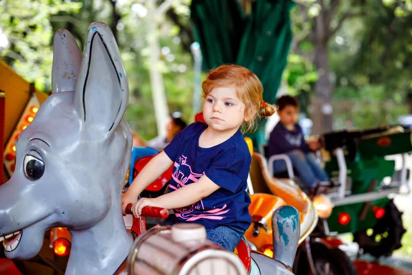 Adorable little toddler girl riding on animal on roundabout carousel in amusement park. Happy healthy baby child having fun outdoors on sunny day. Family weekend or vacations — Stock Photo, Image