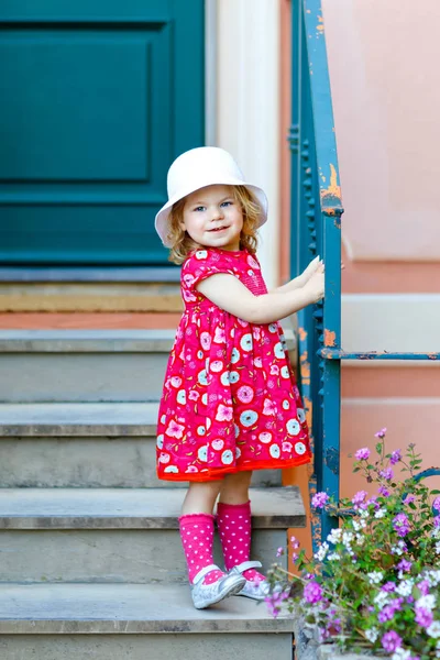 Porträt eines wunderschönen kleinen Mädchens in rosafarbener Sommerkleidung, Modekleid, Kniestrümpfen und Hut. glückliches gesundes Baby posiert vor buntem Haus. — Stockfoto