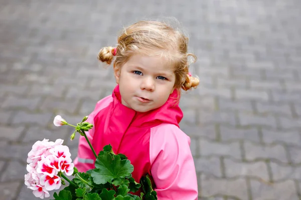 かわいいブロンドの幼児の女の子は、庭にゼラニウムの花の種や苗を植えます。寒い春の日に幸せな健康な赤ちゃん家庭菜園で両親を助ける. — ストック写真