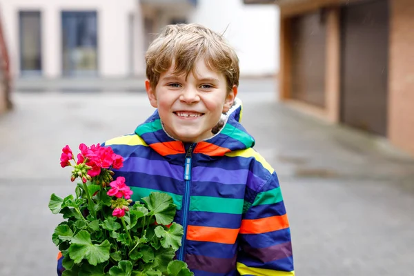 Ragazzo biondo carino scuola piantare semi e piantine di fiori di geranio in giardino. Studentessa che fa progetti di ecologia a scuola. Bambino felice nel freddo giorno di primavera . — Foto Stock