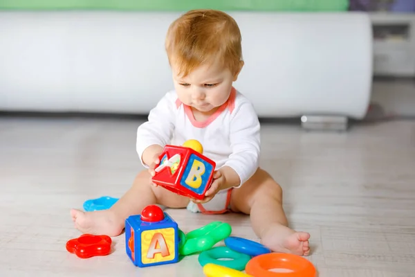 Adorável menina brincando com brinquedos educativos no berçário. Criança saudável feliz se divertindo com brinquedos diferentes coloridos em casa. Criança tentando construir pirâmide de plástico e usando blocos com letras — Fotografia de Stock