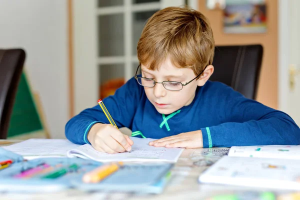 Портрет милого школьника в очках дома, делающего домашнее задание. Маленький сконцентрированный ребенок пишет цветными карандашами в помещении. Начальная школа и образование — стоковое фото