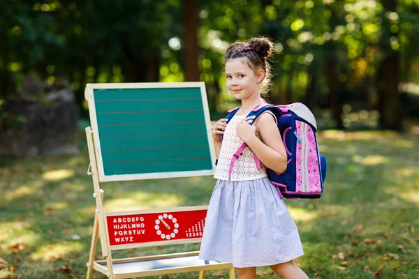 Щаслива маленька дівчинка, що стоїть за столом з рюкзаком або сателітом. Школярка в перший день початкового класу. Повернутися до концепції школи. Здорова чарівна дитина на відкритому повітрі, в зеленому парку — стокове фото