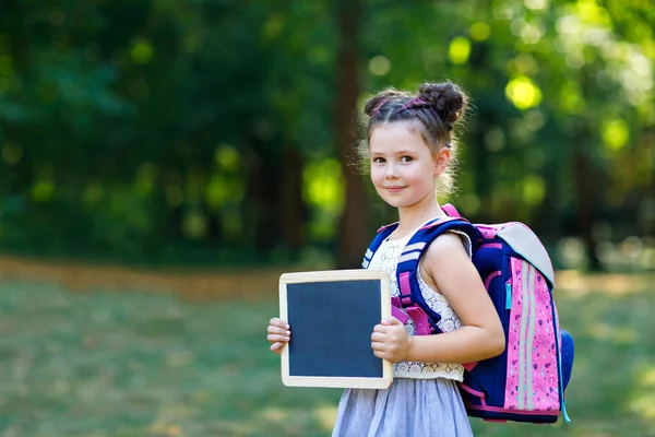 Lycklig liten unge flicka stående med skrivbord och ryggsäck eller skolväska. Schoolkid på första dagen i elementär klass. Friska bedårande barn utomhus, i Green Park. Copyspace tom skrivbord — Stockfoto