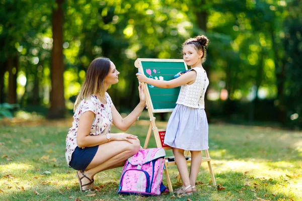 Happy little kid girl and mother by big chalk desk Preschool or schoolkid on first day of elementary class. Back to school concept. Healthy child and woman writing and painting outdoors
