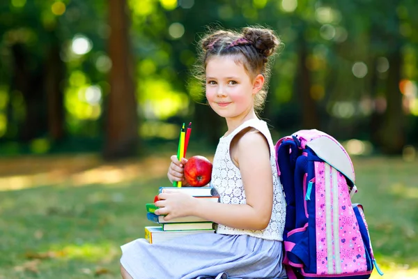 Glücklich entzückendes kleines Mädchen, das am ersten Tag in der Schule oder im Kindergarten Bücher liest und verschiedene bunte Bücher, Äpfel und Bleistifte in der Hand hält. Zurück zum Schulkonzept. Gesundes Kind der Grundschule. — Stockfoto