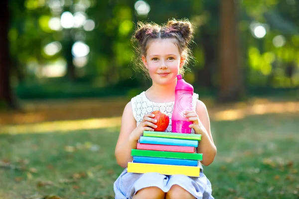 Szczęśliwy adorable kid dziewczynka czytanie książki i gospodarstwa różnych kolorowych książek, jabłka i butelka wody na pierwszego dnia do szkoły lub przedszkola. Powrót do koncepcji szkoły. Zdrowe dziecko elementarne klasy. — Zdjęcie stockowe