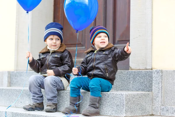 Två barn lite pojkar spelar med blå luft ballonger utomhus. Happy tvillingar och småbarn bröder ler och skrattar tillsammans. Friska barn utanför kall dag i varma kläder. — Stockfoto