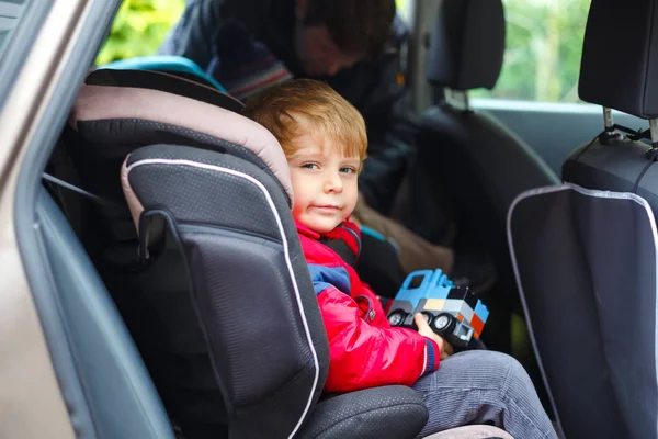 Portrait of pretty toddler boy sitting in car seat. Child transportation safety. Father clips on another son on background. Cute healthy kid boy with toy happy about family vacations with car — Stock Photo, Image