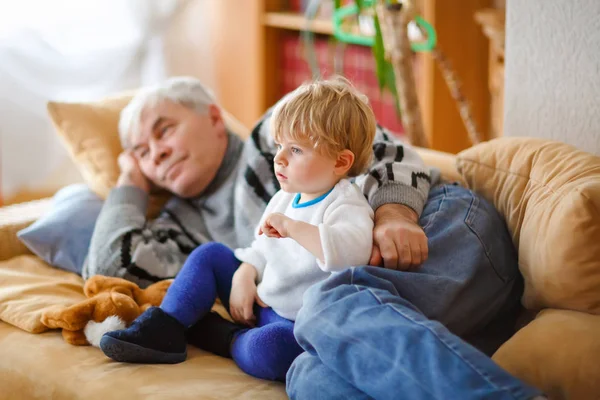 Cute little toddler boy and grandfather watching together tv show. Baby grandson and happy retired senior man sitting together at home with cartoons on television. Calm family time. — Stock Photo, Image
