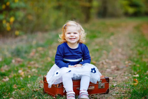 Linda niña pequeña sentada en la maleta en el parque de otoño. Feliz bebé sano disfrutando de caminar con los padres. Día de otoño cálido y soleado con el niño. Ocio activo y actividad con niños en la naturaleza . —  Fotos de Stock