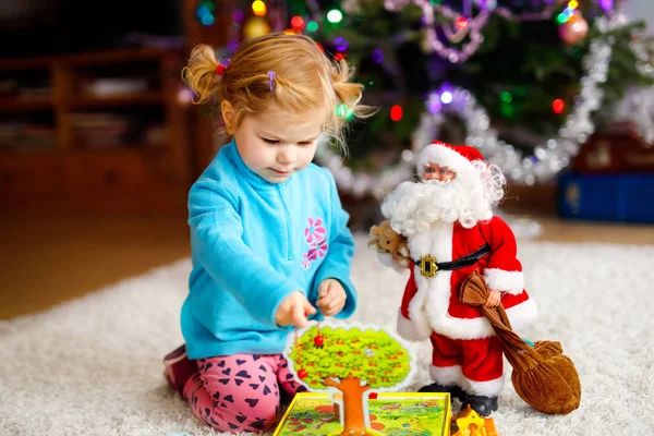 Adorable toddler girl playing with gifts and Christmas Santa Claus toys. Little child having fun with decorated and illuminated Xmas tree with lights on background. Happy healthy funny baby girl. — Stock Photo, Image