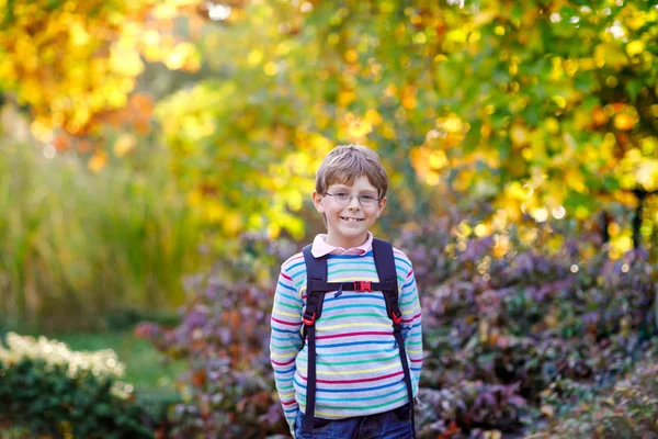 Happy little kid boy with glasses and backpack or satchel on his first day to school on sunny autumn day. Child outdoors with yellow and read maple trees on background, Back to school concept — Stock Photo, Image