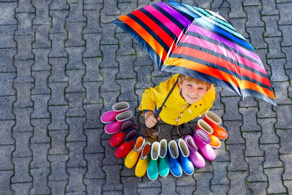 Niño pequeño y grupo de botas de lluvia de colores. Niño rubio de pie bajo el paraguas. Primer plano de los escolares y diferentes botas de goma desde un ángulo alto. Calzado para otoño lluvioso — Foto de Stock