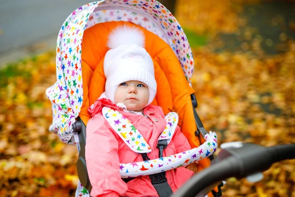 Cute little beautiful baby girl sitting in the pram or stroller on autumn day. Happy healthy child going for a walk on fresh air in warm clothes. Baby with yellow fall maple trees in colorful clothes — Stock Photo, Image