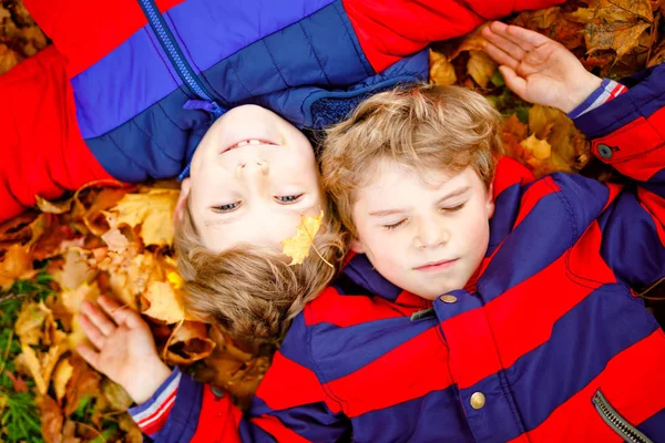 Dos niños pequeños que yacen en las hojas de otoño en ropa de otoño de moda colorida. Felices hermanos sanos divirtiéndose en el parque de otoño en un día cálido. Lindos chicos y mejores amigos sonriendo y riendo — Foto de Stock