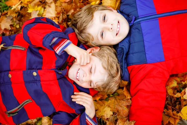 Two little kid boys lying in autumn leaves in colorful fashion fall clothing. Happy healthy siblings having fun in autumn park on warm day. Cute boys and best friends smiling and laughing — Stock Photo, Image