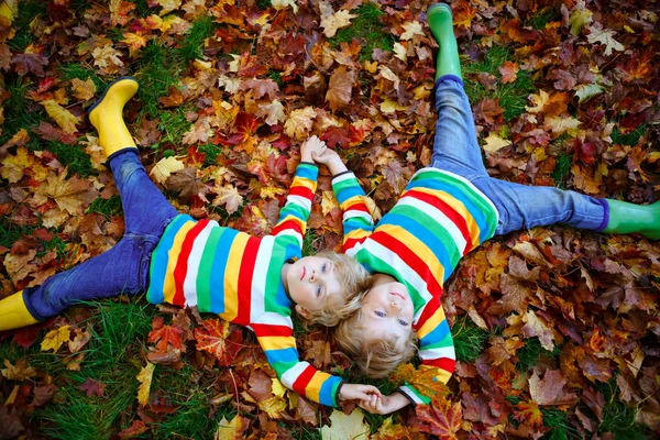Δύο μικρά δίδυμα αγόρια ξαπλωμένα τα φθινοπωρινά φύλλα με πολύχρωμα ρούχα μόδας. Ευτυχισμένα αδέλφια διασκεδάζουν στο πάρκο του φθινοπώρου τη ζεστή μέρα. Υγιή παιδιά με ξανθές τρίχες και μπλε μάτια με φύλλωμα σφενδάμου. — Φωτογραφία Αρχείου