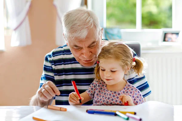 Schattig klein baby peuter meisje en knappe senior grootvader schilderij met kleurrijke potloden thuis. Kleinkind en man hebben plezier samen — Stockfoto
