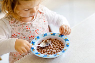 Kahvaltıda sütlü sağlıklı mısır gevreği yiyen yeni yürümeye başlayan kızın yakın ıstabiri. Sevimli mutlu bebek çocuk renkli giysiler içinde mutfakta oturan ve yulaf, tahıl hazırlanması ile eğlenmek. Evde Kapalı