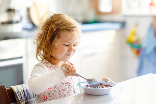 Imádnivaló kislány egészséges gabonapelyhet eszik tejjel reggelire. Aranyos boldog baba színes ruhák ül a konyhában, és szórakozás a felkészülés zab, gabonafélék. Otthon a házban. — Stock Fotó